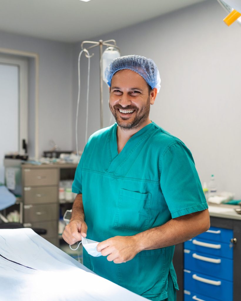 Portrait of male surgeon in operation theater looking at camera showing OK gesture. Doctor in scrubs and medical mask in modern hospital operating room.